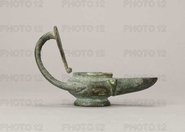 Oil lamp, maker, possibly by Antoni Madeyski 1862–1939 — 1st century, Pozzuoli, oil lamp, weight 0.524 kg., An aged metal vessel with a curving handle and spout sits on a flat surface, showing a weathered, pitted texture and areas of green and brown patina; the body is rounded with a raised central opening and a low pedestal base, the handle rises in a semicircular arc with a small knob at its inner end, and the elongated spout tapers outward, all set against a smooth, neutral beige background.
