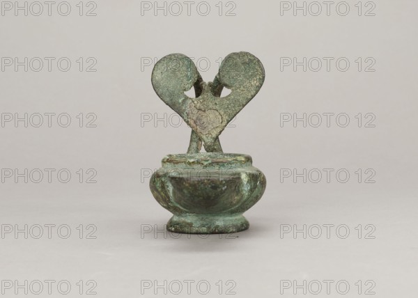 Oil lamp, maker, attributed to Antoni Madeyski 1862–1939 — 1st century; Pozzuoli place of origin ?; oil lamp; weight 0.524 kg., A small corroded metal object sits centered on a neutral light background, showing a rounded bowl-like base topped by a narrow stem and a heart-shaped pair of flat, worn forms joined at the center; the surface displays a mottled patina with shades of green and turquoise, darker green and brown areas, and spots of lighter greenish-gray, with the background appearing pale gray-beige.