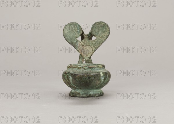 Oil lamp, Madeyski, Antoni 1862–1939, Collection of Ancient Art, Scen.Gal.Class., 1st century, Pozzuoli, oil lamp, weight 0.524 kg, An object with a weathered, textured surface appears centered against a neutral background; it has a rounded bowl-like base rising from a short pedestal and two upward-extending elements that meet near the top to form a heart-like shape with a small gap between them, and the surface shows mottled shades of green and teal with darker green and brown areas and lighter greenish-white spots, all set on a beige-gray backdrop.