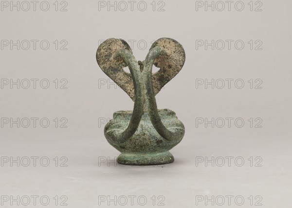 Oil lamp, 1st century, Pozzuoli, oil lamp, weight 0.524 kg, maker name: Madeyski, Antoni 1862–1939, An aged small metal vessel with a rounded base and a short pedestal sits centered on a pale neutral surface against a light gray background; two curved handles rise up from the body and meet above the rim, forming a symmetrical arch that connects to two flat, heart-shaped terminals with rough, pitted surfaces; the vessel and handles show an overall greenish patina with areas of darker green, mottled turquoise, and patches of brown and beige corrosion, while the background and surface are shades of light gray and off-white.