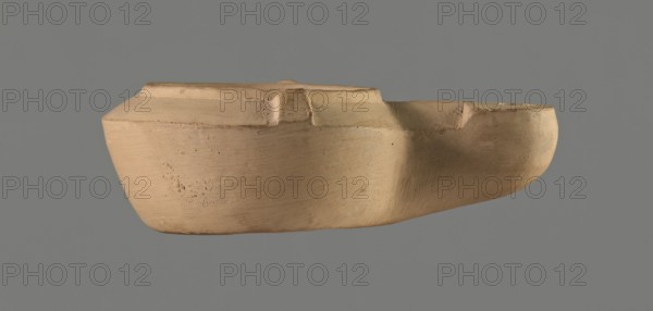 Lamp with comic mask, unknown Italian workshop. Roman oil lamp, Buchi type IX-b = Loeschcke type IXb, from a mould. Round vessel with arms drooping to the outer, separated from the disc by a convex edge; two bars arranged symmetrically on the arms. Concave, flat disc with one olive hole located below a convex theatrical comic mask. Medium-length wick channel decorated with a central groove; rounded burner end. No traces of use no charring. Convex inscription workshop trademark on the bottom: STROBILI. 1st century AD Flavian period, 2nd half of the 18th century, Italy — Magreta, near Modena; Modena. Oil lamp, height 3.4 cm, width 10.1 cm., A single, beige clay object with a smooth, rounded form is shown against a solid gray background; the object has a shallow, open top and an irregular rim with a few raised sections and indentations, a visibly textured surface with subtle horizontal tool marks and small pits, and areas of slightly darker beige shading where the surface curves.