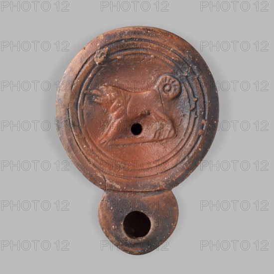 Lamp decorated with a depiction of a dog, unknown eastern workshop, single-burner oil lamp, dogs iconography, 1st century, Roman Empire 27 BC–476, oil lamp, weight 0.082 kg, A round terracotta object with a scalloped rim and a flattened extension at the bottom, showing an embossed animal figure in the central disc. The object displays a range of reddish-brown and dark brown tones with areas of lighter tan and small pale flecks. The central design features a raised depiction of an animal in profile with a curled tail, set within concentric circular grooves; there is a circular hole near the animal's midsection and a larger circular opening in the flattened extension. The surface shows worn and rough texture with small chips and irregularities, and subtle variations in color where the material appears darker around the edges and lighter toward the center.