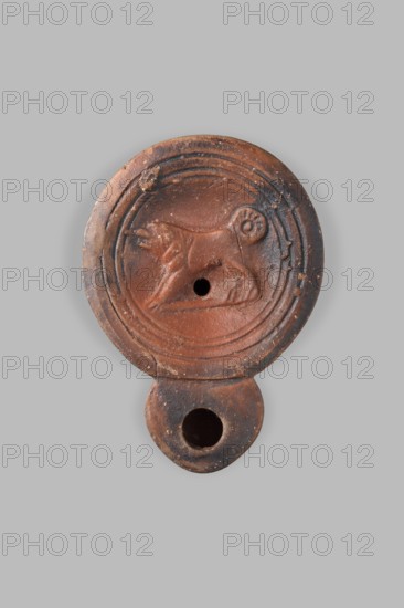 Lamp decorated with a depiction of a dog; unknown eastern workshop; single-burner oil lamp; dogs iconography; 1st century, Roman Empire 27 BC–476; weight 0.082 kg., A round, terracotta object with a molded relief of an animal in profile sits against a light gray background; the object is predominantly reddish-brown with darker brown and blackened areas, showing worn and rough textures and a central hole in the circular field, surrounded by concentric raised rings and a few small chips, and it extends into a smaller rounded projection with another hole, the animal relief featuring a curled tail and a raised head.