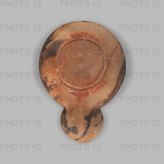 Lamp decorated with a depiction of a dog; unknown eastern workshop; single-burner oil lamp; dogs iconography; 1st century, Roman Empire 27 BC–476; oil lamp; weight 0.082 kg; maker: unknown., A single round, flat object with an attached rounded handle, appearing to be made of clay or terracotta, showing a surface in shades of brown, tan, and reddish-brown with patches of black discoloration and darker burn-like marks; the object has a recessed circular area on its top face and a few small rough spots and speckles in lighter tan and off-white; the background is a uniform light gray.