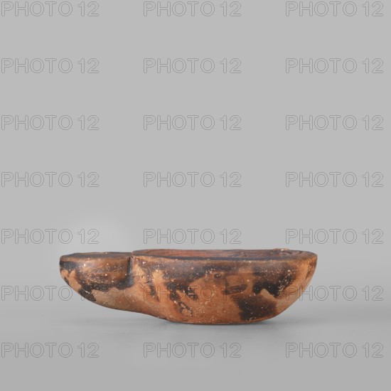 Lamp decorated with a depiction of a dog, unknown eastern workshop, single-burner oil lamp, 1st century, Roman Empire 27 BC–476, weight 0.082 kg, iconography: dogs. Creator: Choynowski, Józef 1833–ca.1915 — collection collector information noted., A shallow, irregularly shaped ceramic bowl sits on a smooth surface against a plain background; the bowl has an elongated form with a rounded end and a slightly raised rim, displaying a mottled surface of earthy brown and black patches with lighter tan flecks and worn areas, and the texture appears rough and weathered with subtle variations in sheen.
