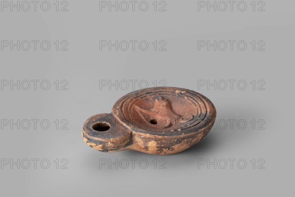 Lamp decorated with a depiction of a dog, unknown eastern workshop; single-burner oil lamp; dogs iconography; 1st century, Roman Empire 27 BC–476; oil lamp; weight 0.082 kg., An earthenware oil lamp with a rounded body and a short handle, shown against a plain gray background; the lamp has a shallow central reservoir with a raised ridged rim and a small circular fill hole, and a slightly protruding nozzle area; the surface shows worn and chipped areas with a patina of dark brown, reddish-brown, and lighter tan tones, and some blackened spots, with textured clay and small flecks of wear visible across the object.