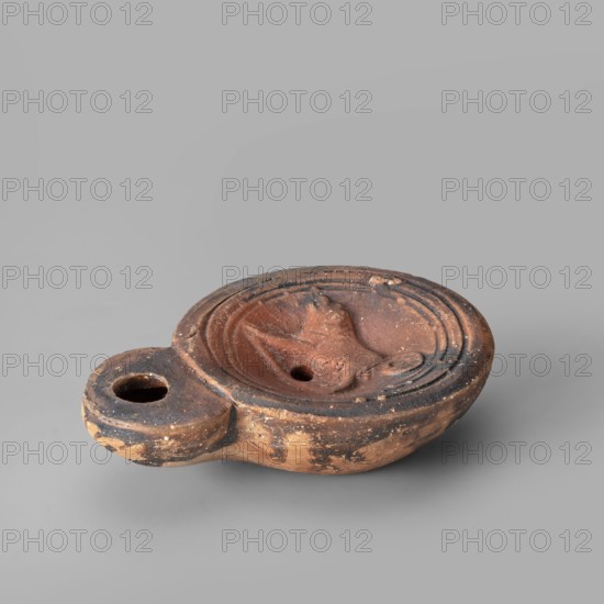 Lamp decorated with a depiction of a dog, unknown eastern workshop, single-burner oil lamp, 1st century, Roman Empire 27 BC–476, weight 0.082 kg, dogs iconography., The image shows an oval-shaped terracotta oil lamp with a rounded handle extension, placed on a smooth gray surface and set against a gray background. The lamp has a central shallow bowl with a small round hole and a molded raised motif in the center, surrounded by a series of concentric ridges. The handle has a circular opening. The surface of the lamp shows worn and rough textures with patches of darker discoloration and small chips. Visible colors include reddish-brown, dark brown, blackened areas, and the surrounding gray background and surface.