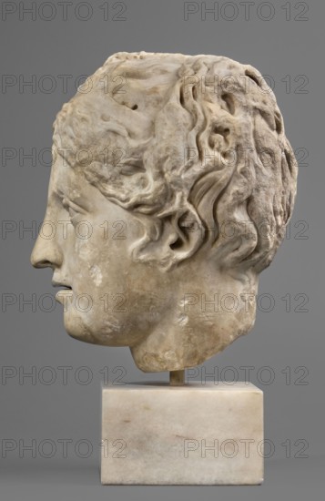 Head of Venus or a nymph, unknown Roman workshop; female portrait, Roman sculpture, hairstyle; Roman copy after a Greek original from the 3rd century BC; early 1st century period of Octavian Augustus, Roman Empire, 27 BC–476; sculpture; height 23 cm, width 16 cm, depth 19 cm, weight 11.13 kg., A carved stone head displayed on a rectangular stone base against a plain gray background, showing a left-facing profile with sculpted hair rendered in flowing carved locks and textured surface; the stone tones include off-white, beige, light tan, and subtle grayish patches, with areas of darker discoloration and surface weathering on the cheek, jaw, and hair; the base is a pale creamy marble with faint beige and gray veining, and a small cylindrical support connects the head to the base.
