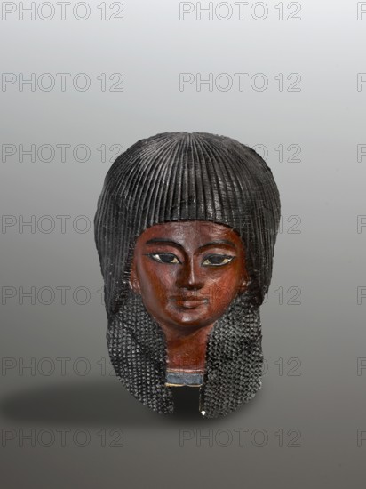 Mummy mask of a boy. The mask of a boy with a delicately modeled, almost girlish face is framed by a black two-layered wig composed of intricately formed curls. Expressive eyes, highlighted with a black line, a slight smile, pierced ears—features of a young man of high birth from the time of Pharaoh Akhenaten or his immediate successors. The upper band of a necklace is visible on the neck. Materials: cartonnage. Type: mask. Face height: 8.5 cm; overall height 24 cm, width 15 cm. Date: c. 1350–c. 1295 BC; New Kingdom, 2nd half 18th Dynasty, Egypt. Iconography: wig, necklace; function: funerary mummy mask., The image shows a carved head with dark brown skin tones and a black textured wig that falls around the sides, the wig featuring vertical ridges on top and a braided or beaded pattern along the sides; the face has black-painted eyes outlined in a lighter color, dark eyebrows, and subtle reddish-brown shading on the cheeks and lips; there is a small area of blue and a thin strip of pal...