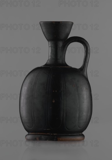 Black-finished aryballoi lekythos, Greek black-glazed pottery lekythos coated with black varnish. Medium size with a flat foot flowing directly into a bulging body, significantly widened in its lower section. Nearly horizontal handles flow into a slender neck ending in a markedly widening, cup-shaped rim. Handle attached at the transition from body to handles and below the cup-shaped rim. Body bears an incised ornament of long, vertically arranged petals. Good condition with minor varnish damage; neck cracked and repaired. 4th century BC, Attica., A dark ceramic jug with a rounded, ribbed body, a narrow neck that flares into a wide mouth, and a single curved handle attached between the neck and shoulder; the surface shows a few small worn spots and scuffs revealing a brownish underlayer, and the overall colors visible are black with touches of brown against a neutral gray background.