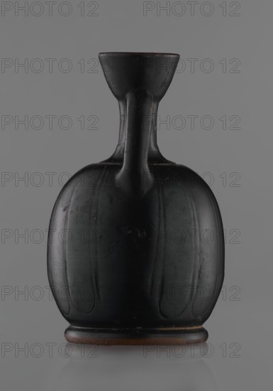 Black-finished aryballoi lekythos, Greek black-glazed pottery. Aryballoi lekythos coated with black varnish. The medium, flat foot flows directly into the bulging body, which is significantly widened in its lower section. The nearly horizontal handles flow into a slender neck ending in a significantly widening, cup-shaped rim. The handle is attached at the transition from the body to the handles and below the cup-shaped rim. The body bears an engraved ornament in the form of long, vertically arranged petals. Good condition, with minor damage to the varnish; the neck is cracked and glued. 4th century BC, Attica, vessel, lekythos., A single dark-colored vessel with a rounded, bulbous body and a narrow neck topped by a flared rim sits centered against a plain gray background; the vessel appears mostly black with subtle darker and lighter areas, a few small brownish spots and a thin brown band at its base, and its surface shows gentle vertical grooves and a smooth, slightly reflective fini...