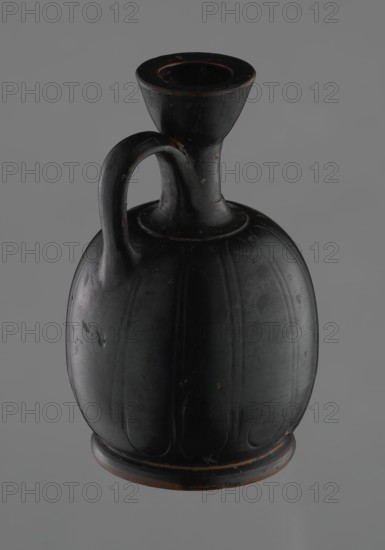 Black-finished aryballoid lekythos coated with black varnish. The medium, flat foot flows directly into the bulging body, which is significantly widened in its lower section. The nearly horizontal arms flow into a slender neck ending in a significantly widening, cup-shaped rim. The handle is attached at the transition from the body to the arms and below the cup-shaped rim. The body bears an engraved ornament of long, vertically arranged petals. Good condition, with minor damage to the varnish; the neck is cracked and glued. 4th century BC, Attic, Greek pottery — vessel lekythos., A single dark vessel with a rounded body and a narrow neck topped by a flared rim, shown against a plain gray background; the vessel has a curved handle connecting the neck to the shoulder and vertical rib-like grooves on its body; the surface shows small scuffs and worn spots revealing a reddish-brown underlayer beneath the dominant black finish; visible colors include black, gray, and reddish-brown.