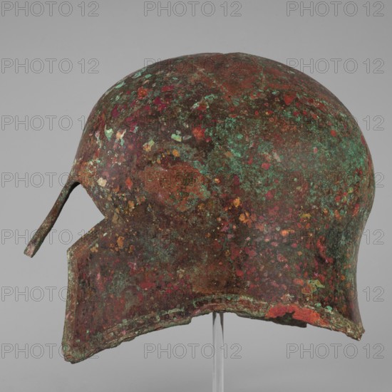 Corinthian-type helmet., Circa 600 BC formerly dated 630–600 BC. Possibly from Sparta or Mycenae according to Otto Schaubert. Bronze helmet with thicker walls in the cheekpieces and a significantly thickened nose plate; domed peak with slightly concave walls widening at the edge. Two small round holes near the back edge for attaching a cord and a similar hole in the peak. Edges of the helmet, cheekpieces, and eyeholes decorated with a precisely crafted band of ornament: rectangular fields framed by double engraved lines and separated by double engraved squares, with rows of tiny applied plastic spheres; an additional row of similar spheres between the top framing lines. Ornamentation reaches the base of the nose guard, which is decorated only with engraved lines echoing the shape of the plate. Function: weapon/helmet. Height 23 cm; width 18.2 cm; depth 25 cm; weight 1.322 kg., An aged metal helmet displayed against a neutral gray background, showing a domed shape with a pronounced chee...
