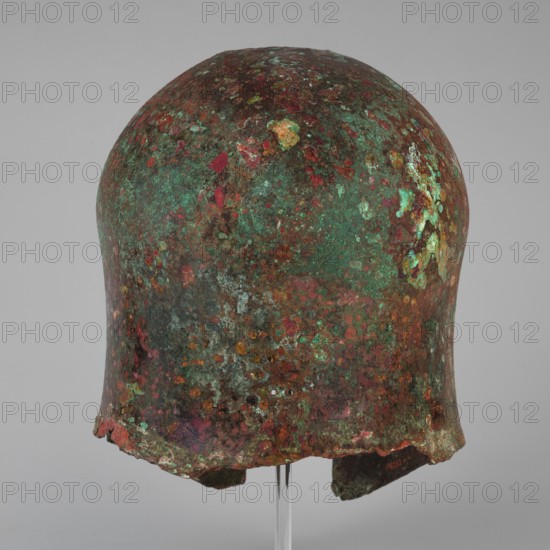 Corinthian-type helmet, This Corinthian-type helmet, with thicker walls in the cheekpieces and a significantly thickened nose plate, has a domed peak, beneath which the walls are slightly concave, widening again at the edge. At the back, near the edge, are two small, round holes for attaching a cord. There is a similar hole in the peak. The edges of the helmet, cheekpieces, and eyeholes are decorated with a precisely crafted band of ornamentation consisting of rectangular fields framed at the top and bottom by a double engraved line and separated by double engraved squares, between which are rows of tiny plastic spheres. A row of similar spheres is located between the lines framing the rectangles at the top. The ornamentation reaches the base of the nose guard, which is decorated only with engraved lines, repeating the shape of the protective plate. Dated ca. 600 BC previously 630–600 BC. Attributed origin: Sparta Greece, — Mycenae Greece per Otto Schaubert. Function: weapon/militaria....