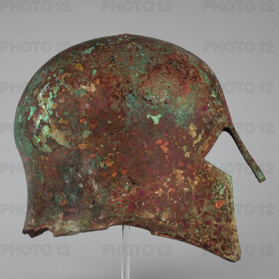 Corinthian-type helmet. This helmet, with thicker walls in the cheekpieces and a significantly thickened nose plate, has a domed peak beneath which the walls are slightly concave, widening again at the edge. At the back, near the edge, are two small round holes for attaching a cord; there is a similar hole in the peak. The edges of the helmet, cheekpieces, and eyeholes are decorated with a precisely crafted band of ornamentation consisting of rectangular fields framed at the top and bottom by a double engraved line and separated by double engraved squares, between which are rows of tiny applied spheres. A row of similar spheres lies between the lines framing the rectangles at the top. The ornamentation reaches the base of the nose guard, which is decorated only with engraved lines repeating the shape of the protective plate. Dated ca. 600 BC previously dated 630–600 BC. Possible origin: Sparta or Mycenae according to Otto Schaubert. Function: weapon. Height 23 cm; width 18.2 cm; depth ...