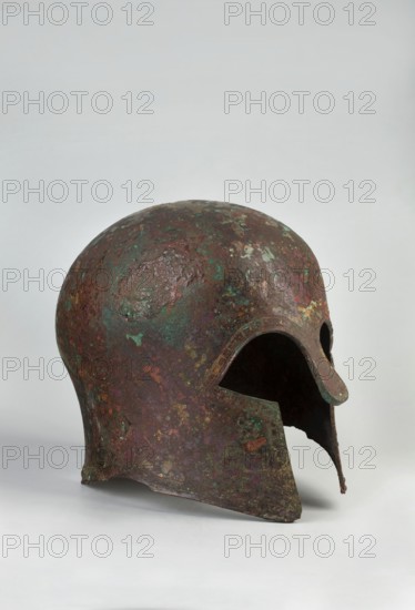 Corinthian-type helmet., Circa 600 BC previously dated 630–600 BC. Possibly Sparta or Mycenae attributions uncertain; Otto Schaubert suggested Mycenae. Bronze helmet with thicker cheekpiece walls and a significantly thickened nose plate; domed peak with slightly concave walls widening at the edge. Two small round holes near the back edge and one in the peak for attaching a cord. Edges of helmet, cheekpieces, and eyeholes decorated with a precise band of ornament: rectangular fields framed by double engraved lines, separated by double engraved squares with rows of tiny applied spheres; an additional row of spheres between the framing lines at the top. Ornamentation reaches the base of the nose guard, which is decorated only with engraved lines repeating the guard’s shape. Function: weapon/militaria. Height 23 cm; width 18.2 cm; depth 25 cm; weight 1.322 kg., A corroded metal helmet with a rounded dome and an open face shows a broken lower edge and a pronounced nose guard, displaying a m...