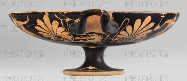 Kylix depicting youths at play, Thalia Painter fl. 520–510 BC. A: three naked youths; B: two youths with spears, one with a rhyton. Palmettes at the vices, lotus buds under the vices. At the bottom, a leaning, naked youth. Greek black-figure ceramic kylix, Attica, dated ca. 510–500 BC. Weight 1.030 kg., A shallow, footed bowl with a rounded base and a short stem sits against a neutral background; the exterior surface shows a black ground with painted decorative motifs in a warm tan color, including stylized palmette or fan-shaped floral elements and curved spiral forms, and there is a looped handle rising from the rim that shares the same black finish with worn tan areas where the surface is chipped, revealing irregular cracks and surface wear; the foot rim shows similar black and tan wear, and the overall palette is primarily black and tan with the neutral gray of the background.