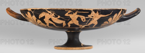 Kylix depicting youths at play, Thalia Painter fl. 520–510 BC. A: three naked youths; B: two youths with spears, one with a rhyton. Palmettes at the vices, lotus buds under the vices. At the bottom, a leaning, naked youth. Greek ceramics, sport in antiquity iconography. Vessel kylix, Attica; 510–500 BC previously dated c. 520 BC. Weight 1.030 kg., A shallow, stemmed bowl with a wide rim and two horizontal handles sits against a neutral background, decorated in contrasting black and orange tones; the exterior band features a scene of human figures rendered in orange on a black ground, showing dynamic poses and flowing drapery and limbs, with a few areas of wear and surface loss revealing lighter orange beneath; decorative palmette-like motifs in orange flank the scene near each handle, and the underside of the bowl and pedestal are predominantly black with some orange showing at the base and rim.