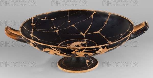Kylix depicting youths at play, Thalia Painter fl. 520–510 BC; scene: A: three naked youths; B: two youths with spears, one holding a rhyton; at the bottom, a leaning naked youth. Decoration includes palmettes at the vices and lotus buds under the vices. Greek ceramic vessel kylix, Attica; dated 510–500 BC previous dating c. 520 BC. Weight 1.030 kg., A shallow, wide bowl with two horizontal handles sits on a low pedestal; the bowl's interior and exterior surfaces show a dark, almost black finish interrupted by a network of lighter, tan-colored cracks and repaired lines, and the rim, handles, and foot display exposed tan areas where the dark surface is worn. The handles are elongated and curved with dark outer surfaces and tan-colored inner faces, and the pedestal foot is dark with tan edges and a visible patch of lighter tone. The interior shows a smooth dark field with irregular tan fissures, while the exterior includes silhouettes and partial figures in the tan color against the dark...