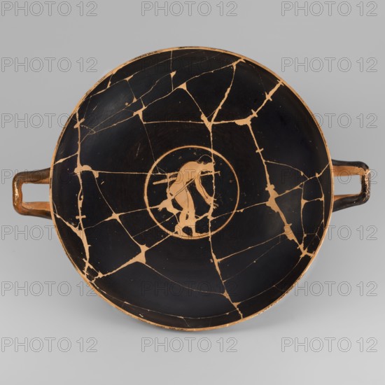 Kylix depicting youths at play, Thalia Painter fl. 520–510 BC. A: three naked youths; B: two youths with spears, one with a rhyton. Palmettes at the handles, lotus buds under the handles. At the bottom, a leaning naked youth. Sport in antiquity iconography; Greek ceramics; vessel kylix. 510–500 BC formerly dated c. 520 BC. Attica. Weight 1.030 kg., A shallow round bowl with two horizontal handles, shown from above, with a black interior and orange-brown lines and areas where the black surface is broken into a network of cracks; at the center a circular orange-brown medallion contains the silhouette of a person in orange-brown against the black, and the handles are orange-brown with worn black areas.