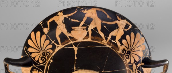 Kylix depicting youths at play, Thalia Painter 520–510 BC. Red-figure kylix, Attica, dated 510–500 BC formerly c. 520 BC. Interior A: three naked youths; exterior B: two youths with spears, one with a rhyton. Palmettes at the handles, lotus buds under the handles; at the bottom, a leaning naked youth. Subject: sport in antiquity. Maker: Thalia Painter fl. 520–510 BC. Vessel, weight 1.030 kg., A fragmentary round ceramic vessel rim with black and terracotta colors shows a painted scene of three nude human figures around a central vessel, with two standing figures reaching toward it and one figure holding a staff; decorative palmette motifs in terracotta appear on either side against the black background, and the piece displays visible craquelure and repaired breaks, with the vessel handles partially visible at the lower edges in black.