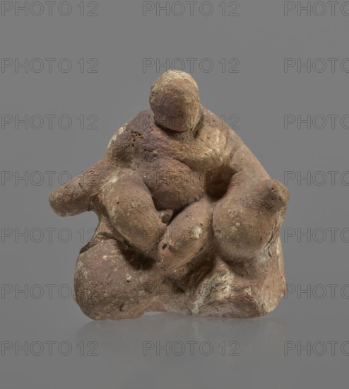 Terracotta figurine of Silenus, Silenus mythology, terracotta. Figurine of an obese Silenus sitting on a rock. The figurine is hollow inside, open at the bottom. The back is only roughly worked, with a large, triangular opening. The clay is red, strongly and unevenly fired; the surface is light, cracked, and mostly chipped. The idol sits frontally, with thighs widely spread and feet crossed. The belly is sagging. The right arm rests with its elbow on the thigh, the forearm and hand stretched slightly downward. The left hand holds a waterskin resting on the rock and the side of the thigh. The small head, slightly lowered, turns to the left and downward, towards the waterskin. Silenus is partially covered by a chlamys, fastened at the right shoulder and thrown back over the left arm; the lower torso and legs are bare. Schematic modeling, head, arms, legs and waterskin rendered synthetically, with a tendency to accentuate volume. Numerous chips and surface flaking. Wide crack in the base ...