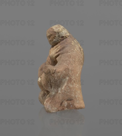 Terracotta figurine of Silenus, Silenus mythology, terracotta. Figurine of an obese Silenus sitting on a rock. The figurine is hollow inside, open at the bottom. The back is only roughly worked, with a large, triangular opening. The clay is red, strongly and unevenly fired; the surface is light, cracked, and mostly chipped. The idol sits frontally, thighs widely spread and feet crossed. The belly is sagging. The right arm rests with its elbow on the thigh, the forearm and hand stretched slightly downward. The left hand holds a waterskin resting on the rock and the side of the thigh. The small head, slightly lowered, turns to the left and downward, towards the waterskin. Silenus is partially covered by a chlamys, fastened at the right shoulder and thrown back over the left arm; the lower torso and legs are bare. Schematic modeling; head, arms, legs and waterskin rendered synthetically, with a tendency to accentuate volume. Numerous chips and surface flaking. Wide crack in the base on th...