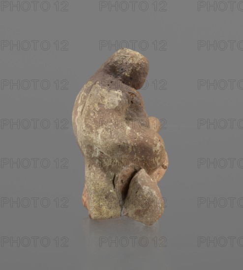 Terracotta figurine of Silenus, Silenus mythology, terracotta. Figurine of an obese Silenus sitting on a rock. The figurine is hollow inside, open at the bottom. The back is only roughly worked, with a large, triangular opening. The clay is red, strongly and unevenly fired; the surface is light, cracked, and mostly chipped. The idol sits frontally, with thighs widely spread and feet crossed. The belly is sagging. The right arm rests with its elbow on the thigh, the forearm and hand stretched slightly downward. The left hand holds a waterskin resting on the rock and the side of the thigh. The small head, slightly lowered, turns to the left and downward, towards the waterskin. Silenus is partially covered by a chlamys, fastened at the right shoulder and thrown back over the left arm; the lower torso and legs are bare. Schematic modeling; head, arms, legs and waterskin rendered synthetically, with a tendency to accentuate volume. Numerous chips and surface flaking. Wide crack in the base ...