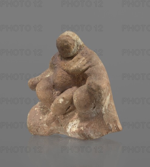 Terracotta figurine of Silenus, Silenus mythology, terracotta. Figurine of an obese Silenus sitting on a rock. The figurine is hollow inside, open at the bottom. The back is only roughly worked, with a large triangular opening. The clay is red, strongly and unevenly fired; the surface is light, cracked, and mostly chipped. The idol sits frontally, thighs widely spread and feet crossed. The belly is sagging. The right arm rests with its elbow on the thigh, the forearm and hand stretched slightly downward. The left hand holds a waterskin resting on the rock and the side of the thigh. The small head, slightly lowered, turns to the left and downward, toward the waterskin. Silenus is partially covered by a chlamys, fastened at the right shoulder and thrown back over the left arm; the lower torso and legs are bare. Schematic modeling; head, arms, legs and waterskin rendered synthetically, with a tendency to accentuate volume. Numerous chips and surface flaking. Wide crack in the base on the ...