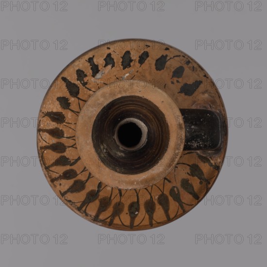 Black-figure lekythos, unknown Attic workshop. Yellowish clay, matte firnia. Disc-shaped foot, ovoid body, slightly sloping handles, wide short neck, rim widening upward. Dark firnia on the rim, outer part of the neck, lower body, and foot. A wide black band below the figural decoration and a narrow black band at the transition from body to handles. Handles decorated with a series of lotus buds connected by intertwining lines; neck with a circular ornament of dots. Belly figural scene: a maenad in a chiton and cloak dancing, facing right with her head turned back; her left arm raised upward and her right bent arm hidden under her cloak. Two dancing satyrs flank her, facing right. Vine branches with grapes in the background. Black-figure pottery, lekythos, c. 500 BC, Attica., A round ceramic object viewed from above with a central cylindrical opening surrounded by a raised rim, showing a terracotta base color with dark brown to black painted leaf-like motifs arranged in a circular patte...