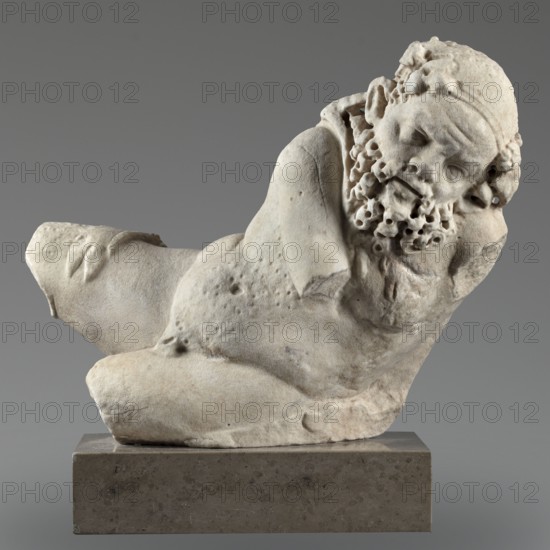 Reclining Silenus, unknown Roman workshop, scenographic gallery class, architectural elements, Silenus mythology, Roman sculpture, sculpture of a reclining Silenus supported on his left hip and elbow, dated 69–79 CE, Roman Empire 27 BC–476, sculpture; height 29.4 cm, width 37 cm, depth 22 cm, weight 19.4 kg, A pale stone sculpture of a reclining male figure with a detailed curly beard and closed eyes rests on a darker rectangular base; the figure shows rough, weathered surface texture with small pits and worn edges, missing parts of the limbs and with a rounded torso and bent arm supporting the head, all set against a smooth gray background, with visible colors including off-white, cream, beige, light tan, gray, and darker brown tones on the base.