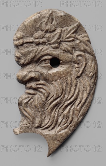 Decoration in the shape of the head of Silenus. Relief appliqué of the head of Silenus, depicted in profile facing left. The nose is wrinkled and snub-nosed with clearly marked nostrils; the lips are full and pursed in a slightly sarcastic grimace. The face is framed by a mustache and beard. The eyes have clearly defined irises, half-closed, with slightly upturned eyebrows. At the level of the lower corner of the eye a round hole is drilled through—remains of a mounting. Behind it, at eye level, a clearly developed, though somewhat schematically depicted, ear is visible. A wreath or crown is visible on the hair above the forehead. A semicircular oval cutout of unknown purpose is visible under the chin. 3rd–2nd century BC, Myrmekion Black Sea region. Appliqué, bas-relief; height 7.2 cm, width 4.1 cm., A carved stone or bone fragment shaped like a stylized human profile with a prominent nose, pronounced brow, and a deep-set circular hole where the eye would be; the profile shows a flowin...