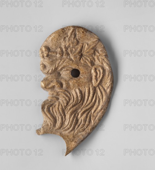 Decoration in the shape of the head of Silenus. Relief appliqué of the head of Silenus depicted in profile facing left. The nose is wrinkled and snub-nosed with clearly marked nostrils; the lips are full and pursed in a slightly sarcastic grimace. The face is framed by a mustache and beard. The eyes have clearly defined irises, half-closed, with slightly upturned eyebrows. At the level of the lower corner of the eye a round hole is drilled through — remains of a mounting. Behind it, at eye level, a clearly developed, though somewhat schematic, ear is visible. A wreath or crown is visible on the hair above the forehead. A semicircular, oval cutout of unknown purpose is visible under the chin. 3rd–2nd century BC, Myrmekion Black Sea region. Appliqué, bas-relief; height 7.2 cm, width 4.1 cm., A carved object shaped like a stylized human profile with a prominent nose, beard, and ear, showing carved flowing hair and leaf-like shapes on the head; the object appears to have a round hole near ...