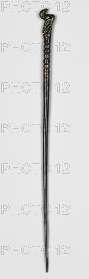 Pin, unknown Luristan workshop. Pin with a thin, oval shaft terminating in a head in the form of a bird figurine. The transition from the shaft to the head begins with a delicately engraved zigzag ornament and parallel lines, behind which is a plastic beaded ornament composed of five segments, alternately spherical with engraved decoration and cylindrical smooth; behind them the extension of the shaft is formed by a figurine of a sitting or swimming bird. Materials: bronze. Subject: birds, geometric ornaments, decorations. Style/period: Luristan bronzes, Iron Period II, 8th–6th century BC 1st half of the 1st millennium BC. Place of origin/discovery: Luristan, Iran. Function: pin. Dimensions: height 21.4 cm; width 0.8 cm. Makers/attributions: unknown Luristan workshop., A long, slender wand-like object with a carved handle shaped like a curved, stylized head and neck, showing carved grooves and ringed bands below the handle leading into a smooth tapered shaft; the object appears glossy ...