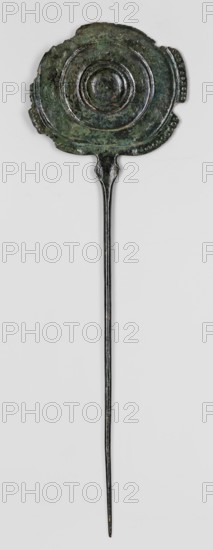 Pin, unknown Luristan workshop. A long pin terminating in a head in the form of an oval shield with geometric decoration. The shaft, circular in cross-section, thickens and widens rhomboidally at the top to form a connector to the shield, which is decorated on one side with punching and engraving. Ornamentation on the shield is arranged concentrically: central round knob with two intersecting engraved bands filled with fine dots; three convex concentric stripes central stripe incised; interwoven braids with dotted interiors in the spaces between stripes; a row of larger dots between the first stripe and the boss; the edge band decorated with a single row of convex dots. The decorative sequence is interrupted at the lower part of the shield where it connects to the pin: on a short section of the edge band the convex dots are replaced by dots, and diagonal incisions appear on the third strip. Engraved geometric ornament also appears on the upper part of the rhomboid connector. Basketry o...