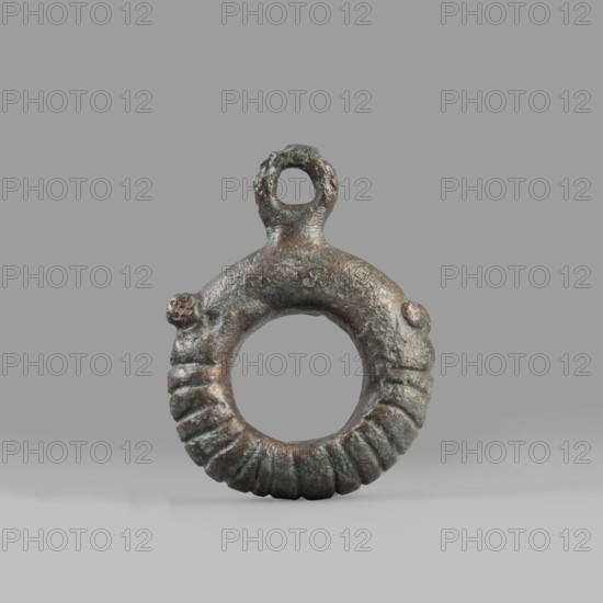 Decorative pendant from a horse harness, unknown Near Eastern workshop. The pendant is in the form of a thick ring with a round eyelet at the top. The lower half of the ring is decorated with 16 notches. On both sides, the notching terminates in a pair of small round knobs. The pendant was probably an element of a harness. 9th–6th century BC end of the 2nd millennium–first half of the 1st millennium BC; Iron Period. Luristan, Near East. Materials: metal; weight 94 g; diameter 5.5 cm; thickness 1.2 cm. Categories: harness components, pendants, decoration, Iranian art., An aged circular metal pendant with a textured, ridged lower half and a rough, mottled upper surface, showing a small loop at the top; the object displays a weathered finish with visible greenish and brownish tones, and is set against a uniform light gray background.