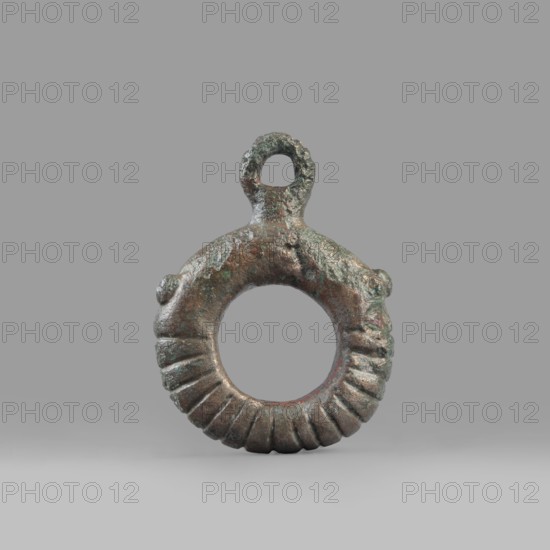 Decorative pendant from a horse harness, unknown Near Eastern workshop. The pendant is in the form of a thick ring with a round eyelet at the top. The lower half of the ring is decorated with 16 notches. On both sides, the notching terminates in a pair of small round knobs. The pendant was probably an element of a harness. 9th–6th century BC end of the 2nd millennium–first half of the 1st millennium BC; Iron Period. Luristan, Near East. Harness component, pendant; material culture; decoration. Weight 94 g; diameter 5.5 cm; thickness 1.2 cm., A circular metal ring with a rough textured surface and a small loop at the top, showing a patina of green and brown with hints of darker gray and reddish tones, the lower half of the ring features a series of carved ridges creating a fluted pattern, the object rests on a smooth light gray background and casts a soft shadow beneath it.