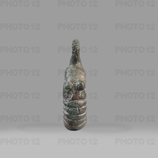 Decorative pendant from a horse harness, unknown Near Eastern workshop. The pendant is in the form of a thick ring with a round eyelet at the top. The lower half of the ring is decorated with 16 notches; on both sides the notching terminates in a pair of small round knobs. The pendant was probably an element of a harness. 9th–6th century BC end of the 2nd millennium–first half of the 1st millennium BC; Iron Period. Provenance: Near East; origin: Luristan. Materials: metal; harness component/pendant; decoration. Weight: 94 g; diameter: 5.5 cm; thickness: 1.2 cm., A small sculptural object stands upright against a plain gray background, showing a textured, weathered surface with a mix of green, brown, and dark bronze tones; the form tapers toward the top with an irregular protrusion, a rounded upper section with two raised rounded areas on the front, and a segmented lower portion with a series of rounded horizontal ridges; lighting casts a soft shadow beneath the object.