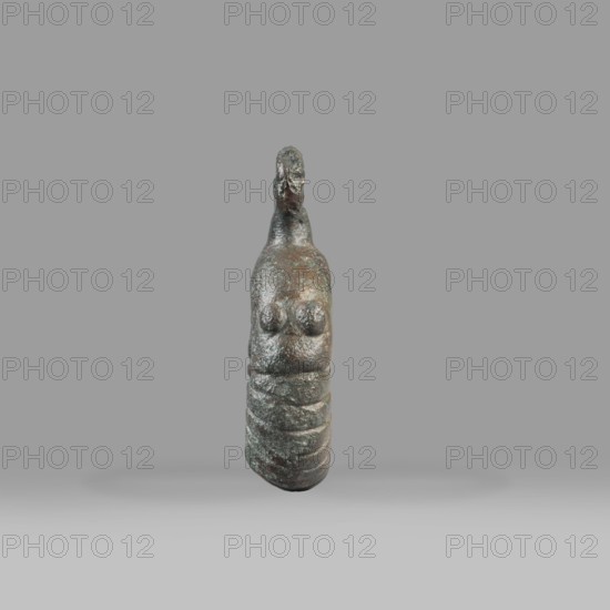 Decorative pendant from a horse harness, unknown Near Eastern workshop. The pendant is in the form of a thick ring with a round eyelet at the top. The lower half of the ring is decorated with 16 notches; on both sides the notching terminates in a pair of small round knobs. The pendant was probably an element of a harness. Iron Period, 9th–6th century BC end of the 2nd millennium–first half of the 1st millennium BC. Luristan, Near East. Materials: metal; weight 94 g; diameter 5.5 cm; thickness 1.2 cm., A small sculpted figure with a rounded, elongated form standing upright against a plain gray background, showing a rough, weathered surface with visible textural irregularities; the figure has a narrowed top that tapers into a small head-like shape and a broader midsection with two raised, rounded protrusions on the front, followed by a segmented lower portion with horizontal grooves; colors visible include dark brown, greenish patina, and hints of black and gray across the surface, with ...
