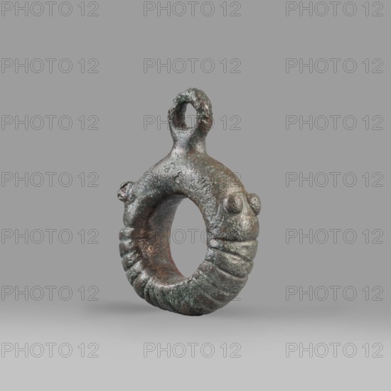 Decorative pendant from a horse harness, unknown Near Eastern workshop. The pendant is in the form of a thick ring with a round eyelet at the top. The lower half of the ring is decorated with 16 notches; on both sides the notching terminates in a pair of small round knobs. The pendant was probably an element of a harness. Iron Period, 9th–6th century BC end of the 2nd millennium–first half of the 1st millennium BC. Luristan, Near East. Materials: unspecified metal, weight 94 g, diameter 5.5 cm, thickness 1.2 cm., An irregular ring-shaped metal object with a rough, pitted surface and a looped extension at the top, resembling a small pendant or amulet; the ring has a textured, segmented lower half with raised ridges and a pair of small rounded protrusions on one side that look like stylized eyes, and a small rounded knob on the opposite side; the interior of the ring is hollow and shows uneven surfaces; the object rests against a smooth, neutral background. Colors visible include greenis...