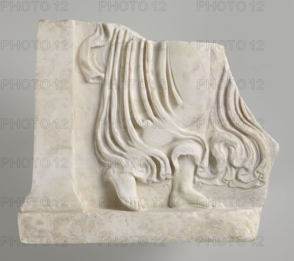 Fragment of a relief with a dancing maenad, 1st century BC, 27 BC–14 AD period of the rule of Octavian Augustus, Roman Empire, bas-relief, height 35 cm, width 30 cm, thickness 6.5 cm, A carved marble fragment showing the lower part of a draped figure with a few visible feet and flowing folds of cloth, all rendered in pale off-white and creamy beige tones with subtle areas of light gray and faint warm discoloration; the drapery forms several rounded pleats and curling edges, and the feet rest on a rectangular base of the same stone; the background is a smooth neutral gray.