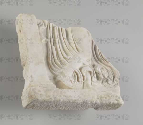 Fragment of a relief with a dancing maenad, 1st century BC 27 BC–14 AD, period of the rule of Octavian Augustus, Roman Empire, bas-relief; height 35 cm, width 30 cm, thickness 6.5 cm, A weathered carved stone fragment rests against a neutral gray background, showing a draped, folded fabric motif with flowing curved grooves and rounded, rolled forms. The stone surface displays a range of light colors including off-white, creamy beige, pale yellowish tones, and faint warm browns in spots, with subtle shadows in gray and soft tan where the carving recesses. The piece has rougher, eroded edges and a textured underside, while the carved areas appear smoother with gently rounded contours and layered ridges suggesting folds.