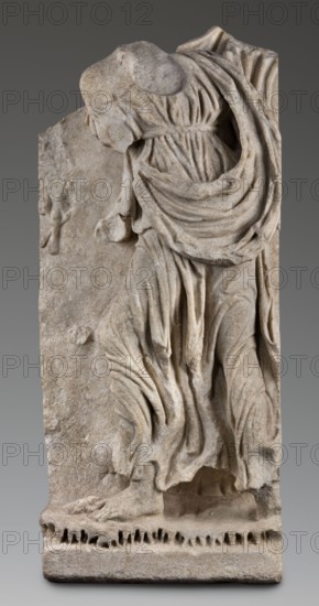 Fragment of a sarcophagus depicting the daughter of King Lycomedes, unknown Roman workshop, classified Gallic scenography, Lycomedes mythology, Roman art, late 2nd–3rd century, Roman Empire 27 BC–476, bas-relief, sarcophagus Roman, height 77 cm, width 32.5 cm, depth 16 cm, weight 63.5 kg, A partially preserved stone relief of a draped figure with visible folds and flowing fabric in shades of beige and light gray against a darker gray background; the figure's head and parts of limbs are missing or worn, and the carved base shows irregular perforations and rough texture in similar beige-gray tones.