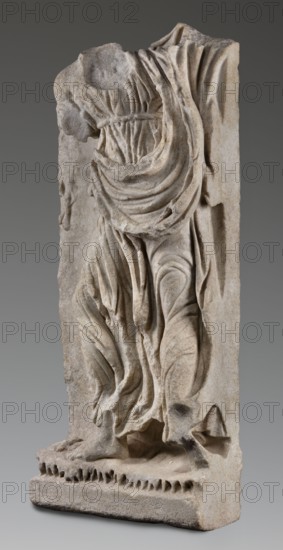 Fragment of a sarcophagus depicting the daughter of King Lycomedes, unknown Roman workshop, Classified Gallic Scenography, Lycomedes mythology, Roman art, late 2nd–3rd century, Roman Empire 27 BC–476, bas-relief sarcophagus, height 77 cm, width 32.5 cm, depth 16 cm, weight 63.5 kg, A carved stone relief fragment showing the draped torso and lower body of a figure with flowing, layered garments, displayed against a neutral gray background; the stone has a weathered, textured surface in shades of beige, off-white, and light gray, with shadows accentuating the folds and carved details, and a darker gray area near the lower right where the stone appears more worn.