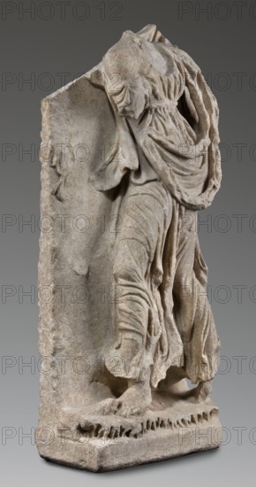 Fragment of a sarcophagus depicting the daughter of King Lycomedes, unknown Roman workshop, Classified Gallic Scenography, Lycomedes mythology, Roman art, late 2nd–3rd century, Roman Empire 27 BC–476, bas-relief sarcophagus, height 77 cm, width 32.5 cm, depth 16 cm, weight 63.5 kg, A carved stone sculpture fragment showing the lower torso and legs of a draped figure set against a rectangular stone backing; the figure wears layered, flowing garments with deep, rounded folds and gathered fabric around the waist and shoulders, and the feet rest on a textured base. Visible colors include light beige, gray, and warm tan tones in the stone, with subtle darker gray shading in recessed areas. The surface shows rough and worn textures, with smooth, sculpted drapery contrasts and some chipped and eroded edges along the backing and base.