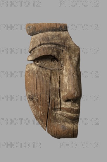 Fragment of a sarcophagus mask. Preserved half of a face made from a single piece of wood. Above the low forehead is a preserved fragment of a wig; below is a recess with remnants of adhesive where an eyebrow made of a different material was probably attached. Almond-shaped eye with preserved eye socket and thick adhesive layer where an inlaid eye was placed. Long, slender nose preserved except for the left cover of the nostril. Mouth with thin lips and slightly upturned corners. Traces of mortar preserved throughout, with minimal remnants of beige paint. On the reverse a stepped niche is carved at mouth level, also showing traces of mortar. The chin has space for a beard. A knot is present on the right cheek. Face height: 17 cm. c. 1400–c. 1295 BC; New Kingdom, late 18th Dynasty decline. Findspot: Deir el-Medina. Sarcophagus mask; overall height 18.5 cm, width 9 cm, depth 4.5 cm., A carved wooden fragment of a human face shown against a plain gray background, with the wood displaying ...