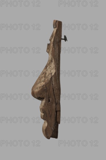 Fragment of a sarcophagus mask. Preserved half of a face made from a single piece of wood. Above the low forehead is a preserved fragment of a wig; below is a recess with remnants of adhesive, where an eyebrow made of a different material was probably attached. Almond-shaped eye; preserved eye socket contains a thick layer of adhesive that once secured an inlaid eye of different material. Long, slender nose preserved in its entirety except for the left nostril cover. Mouth with thin lips and slightly upturned corners. Traces of mortar throughout, with minimal remnants of beige paint. On the reverse, a stepped niche is carved at mouth level, also showing traces of mortar. Chin has space for a beard. A knot is present on the right cheek. Face height: 17 cm. Date: c. 1400–c. 1295 BC, New Kingdom, late 18th Dynasty decline. Find location: Deir el-Medina. Object type: sarcophagus mask. Dimensions: height 18.5 cm, width 9 cm, depth 4.5 cm., A vertical piece of weathered wood with a rough, ca...