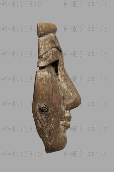 Fragment of a sarcophagus mask. Preserved half of a face made from a single piece of wood. Above the low forehead is a preserved fragment of a wig; below is a recess with remnants of adhesive where an eyebrow made of a different material was probably attached. Almond-shaped eye with preserved eye socket containing a thick layer of adhesive that once held an inlaid eye of different material. Long, slender nose preserved in its entirety except for the left cover of the nostril. Mouth with thin lips and slightly upturned corners. Traces of mortar preserved throughout, with minimal remnants of beige paint. On the reverse, a stepped niche is carved at mouth level, also showing traces of mortar. The chin has space for a beard. A knot is present on the right cheek. Face height: 17 cm. c. 1400–c. 1295 BC; New Kingdom, late 18th Dynasty decline; findspot: Deir el-Medina. Sarcophagus mask; overall height 18.5 cm, width 9 cm, depth 4.5 cm., A weathered wooden mask shaped like a human profile with...