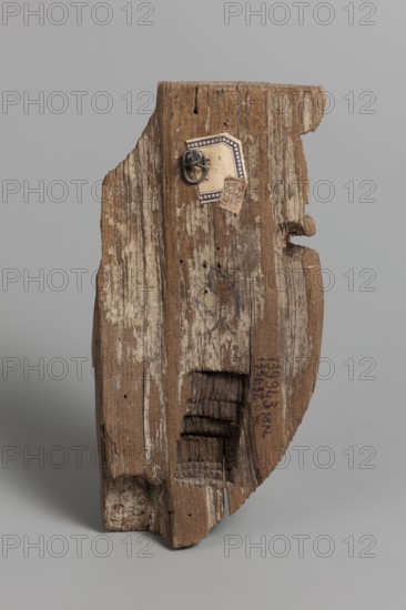 Fragment of a sarcophagus mask. Preserved half of a face made from a single piece of wood. Above the low forehead is a preserved fragment of a wig; below is a recess with remnants of adhesive, in which an eyebrow made of a different material was probably attached. Almond-shaped eye, preserved eye socket with a thick layer of adhesive, in which an eye made of a different material was placed. Long, slender nose preserved in its entirety, except for the left cover of the nostril. Mouth with thin lips and slightly upturned corners. Traces of mortar preserved throughout, with minimal remnants of beige paint. On the reverse, a stepped niche is carved at mouth level, also showing traces of mortar. The chin has space for a beard. A knot is present on the right cheek. Face height: 17 cm. c. 1400–c. 1295 BC; New Kingdom, late 18th Dynasty decline. Find location: Deir el-Medina. Sarcophagus mask; height 18.5 cm, width 9 cm, depth 4.5 cm., A weathered wooden plank with rough, uneven edges and a ve...