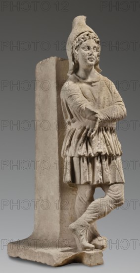 Fragment of a monopodium, unknown Roman workshop, restitution Lubusz Land, Lubniewice Lubusz Voivodeship — furniture, Roman sculpture, Attys myth., 2nd century, Roman Empire 27 BC–476, sculpture; height 77 cm, width 31 cm, depth 34 cm, weight 64 kg, A pale beige stone statue of a standing figure leaning against a rectangular support, wearing a pointed hat and layered, pleated clothing with long sleeves and a skirt-like lower garment; the figure has folded arms, crossed legs with wrapped lower legs and footwear, and a detailed carved face with stylized hair. The background is a smooth gradient of gray, and the statue shows subtle weathered texture and roughness in off-white and light tan tones.